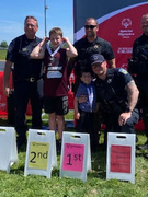 A student in a maroon jersey holds his medal while standing next to another young student and three smiling police officers. They are posing outdoors in front of a red Special Olympics Illinois banner and podium signs marked 1st and 2nd.